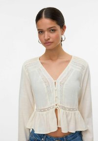 Krämfärgad blus med peplum-nederkant, knappstängning och spetsdetaljer längs halsringningen och ärmarna. Långa ärmar med mjuk textur.