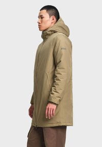 Homme portant un manteau d'hiver beige à capuche, avec fermeture éclair à l'avant et poches latérales, debout de profil sur un fond uni.