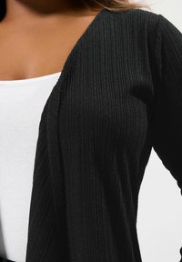 Svart ribbad cardigan med V-halsdesign, med en mjuk textur och draperad framsida. Lager på en vit tanktop.