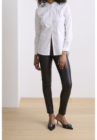 Camisa de botón de color blanco con cuello, combinada con pantalones de cuero sintético negros y mules de tacón negro con punta, que presentan un detalle decorativo de hebilla.