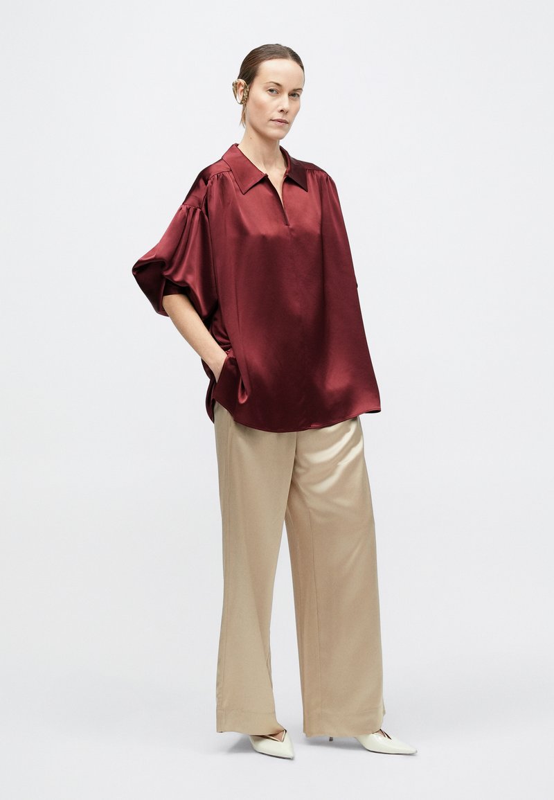 Woman standing with hands in pockets wearing a loose maroon satin blouse, wide beige pants, white pointed shoes, and a hearing aid.