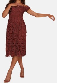 Tummanpunainen pitsimekko olkapäät paljastavilla hihoilla; kerroksellinen muotoilu, jossa on yksityiskohtaisia kukkaleikkauksia ja polven mittainen helma. Nilkkaremmit ja beiget korot.