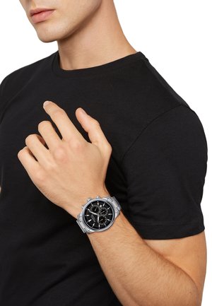 Montre chronographe argentée avec un cadran noir, dotée de trois sous-cadrans, d'un bracelet en métal et d'un boîtier rond. Portée sur un poignet à la teinte de peau claire.