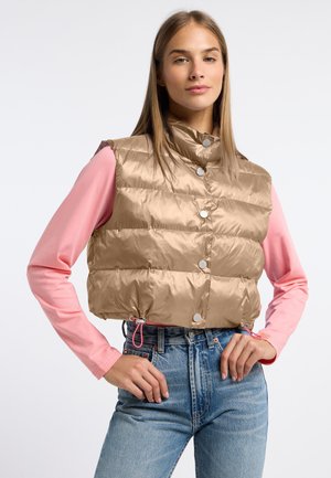 Młoda kobieta w beżowej, krótko skrojonej pikowanej kamizelce z zatrzaskami, nosząca różową bluzkę z długim rękawem oraz wysokim stanem niebieskie dżinsy.