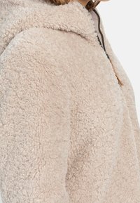 Hoodie en polaire beige avec un extérieur texturé et doux, doté d'une fermeture éclair à l'avant et d'un léger col, conçu pour la chaleur et le confort.