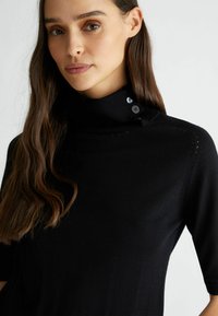 Maglione nero a collo alto con maniche corte, caratterizzato da dettagli con bottoni e una texture liscia. Cuciture sottili lungo le spalle.