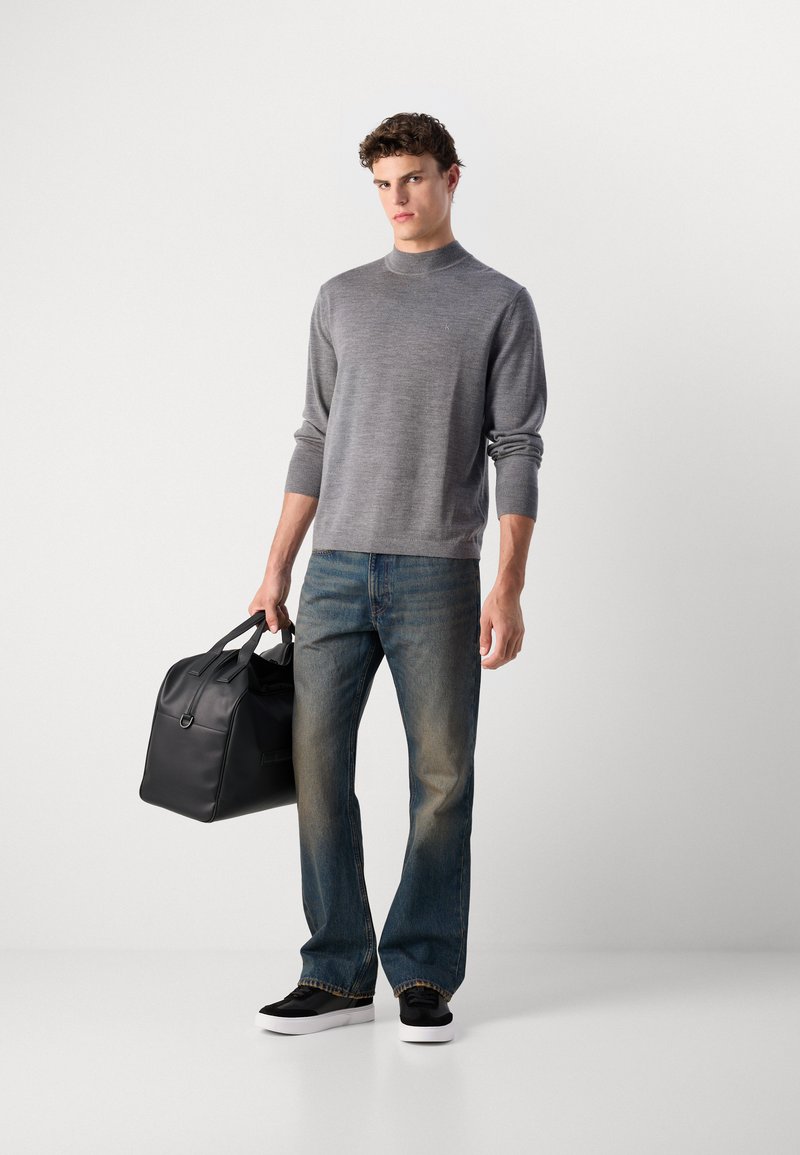 Grauer Strickpullover mit hohem Kragen, kombiniert mit blauen Jeans. Eine schwarze Ledertasche und schwarze Schuhe runden das Outfit ab.