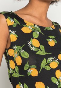 Nära bild av en ärmlös svart klänning med tryck av gula citroner och gröna blad, bärs av en person mot en enhetlig bakgrund.