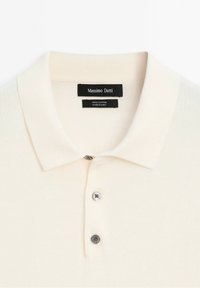 Polo en coton de couleur beige clair avec un col plat, une patte de boutonnage à trois boutons et une texture lisse. L'étiquette indique "Massimo Dutti, 100 % coton."