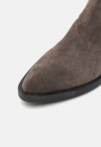 Brązowe zamszowe botki za kostkę z ostrym noskiem i czarną gumową podeszwą. Tekstura jest gładka, prezentująca miękki, bogaty materiał i czysty design.