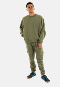 Conjunto de sudadera y pantalones de chándal en verde oliva con un corte relajado; presenta puños acanalados, textura suave y sutiles detalles laterales. Combinado con zapatillas azules.