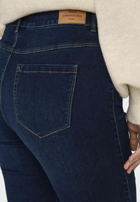 Mörkblå denimjeans med hög midja. Har en ljusbrun läderlapp på baksidan och en enda bakficka med detaljer i gul söm.