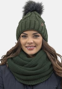 Gorra de punto verde con un pompón negro esponjoso en la parte superior, combinada con una bufanda infinita verde texturizada a juego. Ambas están hechas de hilo suave.