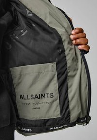 Užtrauktuku užsegamas švarkas turi alyvuogių žalumyną su juodu vidumi. Pridedama išorinė kišenė, pažymėta „ALLSAINTS“, su tekstu skirtingomis kalbomis.