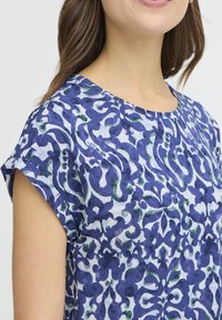 Sinine ja valge mustriga pluus lühikeste varrukatega, ümmarguse kaelusega ning abstraktsete lillemotiividega erinevates sinise ja rohelise toonides.