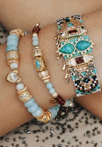 Trois bracelets sur un poignet : un bracelet en perles d'or et de turquoise, un bracelet en perles multicolores dorées et un bracelet tressé avec des pierres précieuses.