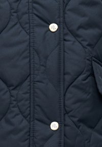 Μπλε ναυτικό καπιτονέ μπουφάν με διαγώνια ραφή, χρυσά μεταλλικά στοιχεία και μια κουμπί με λογότυπο πάνω σε λεία υφασμάτινη επιφάνεια.