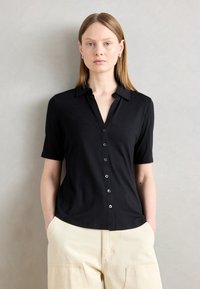 Camicia nera a maniche corte con colletto, caratterizzata da una texture liscia e chiusura frontale con bottoni. Abbinata a pantaloni color crema.