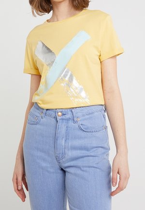 Mujer vistiendo una camiseta amarilla con un diseño de X en pinceladas plateadas y azul claro, combinada con jeans de tiro alto en azul claro.