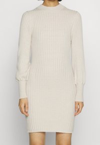 Beżowa dzianinowa sukienka z długim rękawem i wysokim kołnierzem. Teksturowana tkanina, dopasowany krój oraz marszczone mankiety przy nadgarstkach.