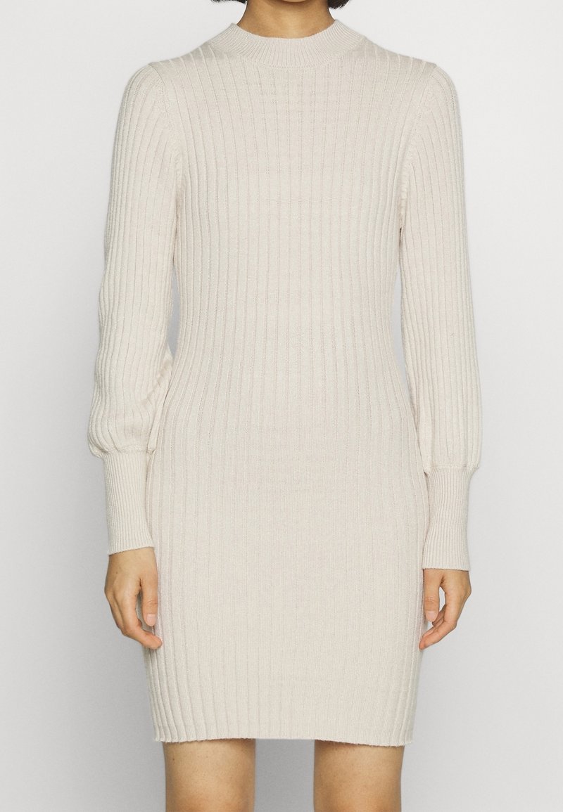 Beżowa dzianinowa sukienka z długim rękawem i wysokim kołnierzem. Teksturowana tkanina, dopasowany krój oraz marszczone mankiety przy nadgarstkach.
