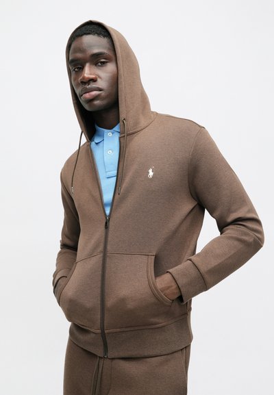 Smeđa dukserica na rajsferšlus s prednjim džepom, s malim bijelim logotipom na prsima i kapuljačom. Nosimo je preko plave polo majice.