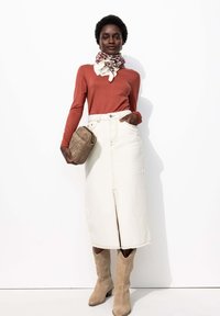 Haut à manches longues rouille associé à une jupe en jean blanche avec une fente à l'avant. Les accessoires comprennent une écharpe à motif et un sac marron texturé.