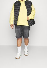 Gilet nero imbottito sopra una felpa gialla con cappuccio, abbinato a pantaloni corti in denim scuro e sneakers bianche con dettagli verdi. Sono indossati calzini bianchi alla caviglia.