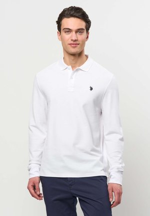 Mann trägt weißes langärmeliges Poloshirt mit schwarzem Logo auf der Brust und dunkelblaue Hose, steht vor weißem Hintergrund.