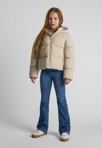 Giacca imbottita beige con cappuccio, dotata di chiusura a zip e maniche ampie. Abbinata a jeans viola svasati e scarpe da ginnastica bianche.