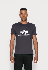 Tmavě šedé bavlněné tričko s bílým logem a emblémem a textem "ALPHA INDUSTRIES" umístěným uprostřed na přední straně. Krátké rukávy, kulatý výstřih.