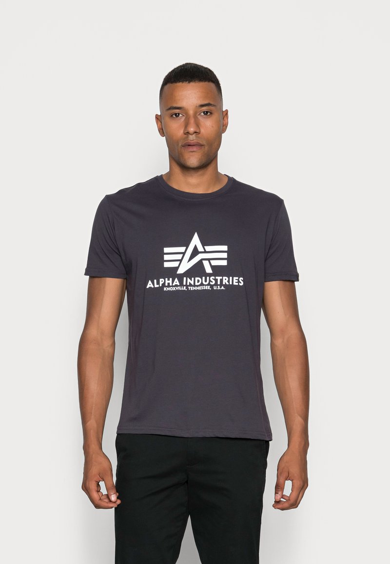 Tmavě šedé bavlněné tričko s bílým logem a emblémem a textem "ALPHA INDUSTRIES" umístěným uprostřed na přední straně. Krátké rukávy, kulatý výstřih.