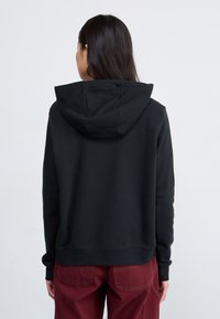 Sweatshirt preto com capuz, de corte descontraído, textura suave e punhos canelados. Usado com calças bordô, mostrando uma vista de costas casual.
