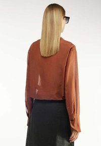Rostfarbige, transparente Bluse mit langen Ärmeln, geknöpften Manschetten und lockerer Passform, kombiniert mit einem schwarzen Rock. Glatte Textur und minimalistisches Design.