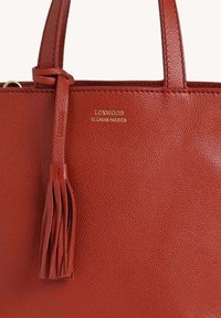 Sac à main en cuir rouge avec un détail de houppes et un logo embossé doré "Loxwood Le Carris Parisien" sous les poignées.