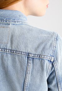 Ljust blå denimjacka med klassisk krage, synlig sömnad och en strukturerad tyg, som visar ryggen och axelområdet.