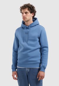 Jongeman met krullend haar in een lichtblauwe hoodie met de tekst "PURE PATH" en blauwe jeans, die iets opzij kijkt tegen een effen achtergrond.