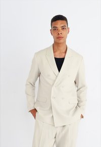 Blazer beige de doble botonadura con con solapas de muesca, dos bolsillos frontales y textura suave, llevado sobre una camisa negra. Ajuste personalizado, diseño minimalista.