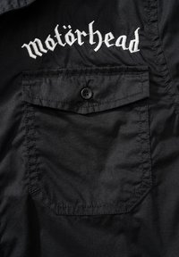 Juodas marškinėliai su priekinėmis kišenėmis ir balta siuvinėta "motörhead" logotipu krūtinėje, pagaminti iš lygaus, švelnaus audinio.