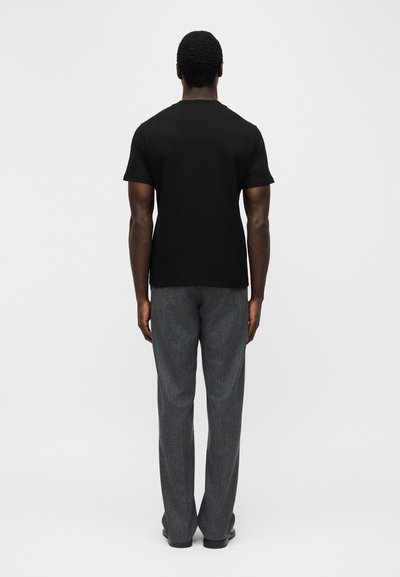Άνδρας που στέκεται γυρισμένος, φορά απλό μαύρο κοντομάνικο μπλουζάκι και γκρι παντελόνι ίσιας γραμμής, μπροστά σε λευκό φόντο.