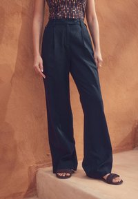 Sort, vid, højtaljede bukser med folder foran, lavet af let stof; kombineret med mørke flade sandaler. Baggrunden er struktureret.