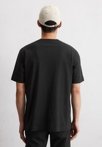 Zwart T-shirt met korte mouwen van katoen, met een ronde hals en een ontspannen pasvorm. Een beige basebalpet wordt gedragen, met een verstelbare band aan de achterkant.