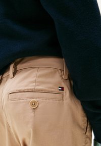 Pantaloni beige con tasca sul retro chiusa da bottone e piccolo logo rosso, bianco e blu, abbinati a un maglione blu scuro.