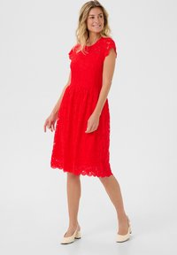 Rochie roșie din dantelă, cu un corset ajustat și o fustă evazată. Mânici scurte tip capă și tiv crestat, asortată cu pantofi cu toc bloc bej.