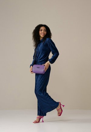 Marineblauwe geplooide jurk met lange mouwen, gecombineerd met een paarse crossbodytas en roze sleehakken. Gladde textuur, ontspannen pasvorm, elegante uitstraling.