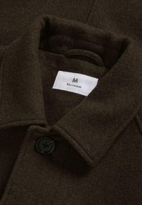 Tamno smeđi vuneni kaput sa struktuiranim kragnom, jednim detaljem crnog dugmeta i bijelom etiketom s natpisom "Matinique". Mekana tekstura.