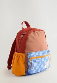 Zainetto piccolo con parte superiore arancione bruciato, davanti color pelle, tasche laterali gialle e tasca frontale con cerniera a quadretti blu e cursore arancione.