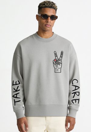 Mann trägt grauen Pullover mit Friedenshand-Grafik und den Worten Glaube, Liebe, Frieden, Freude, Hoffnung; "TAKE" am linken Ärmel und "CARE" am rechten Ärmel.