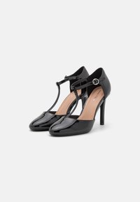 Un paio di scarpe con tacco alto a T in pelle verniciata nera, con punta arrotondata e piccola fibbia argentata su sfondo bianco.