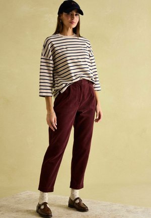 Gestreiftes creme- und marineblaues Langarmhemd, burgunderfarbene Cordhosen, weiße Socken mit Punkten und braune Slipper, kombiniert mit einer schwarzen Baseballkappe.